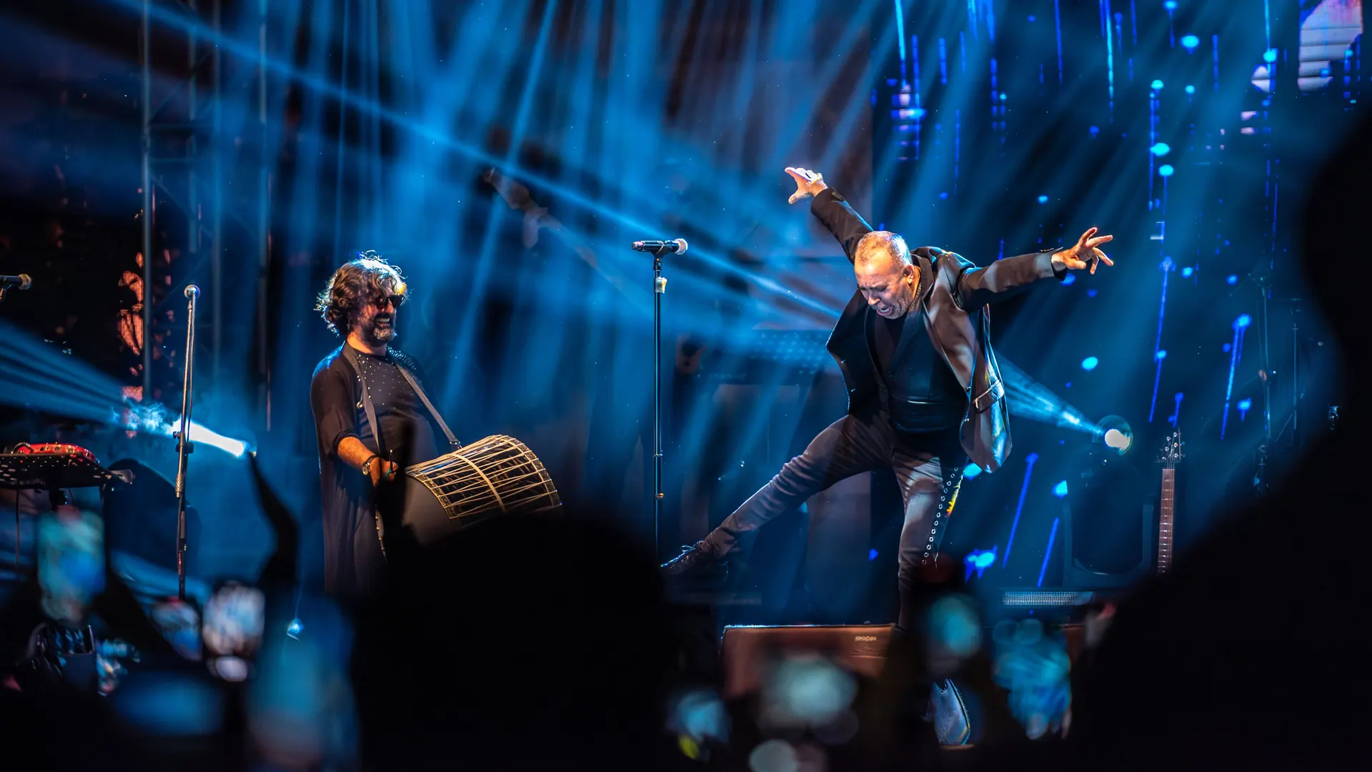 A dancer leaps with arms spread wide in a zeybek on a stage lit by blue beams, while a drummer with a davul stands nearby.